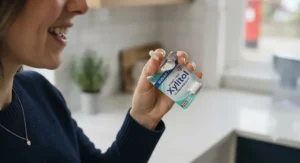 A friendly UK dentist in a modern clinic smiling and holding up a pack of sugar-free gum to discuss oral hygiene.